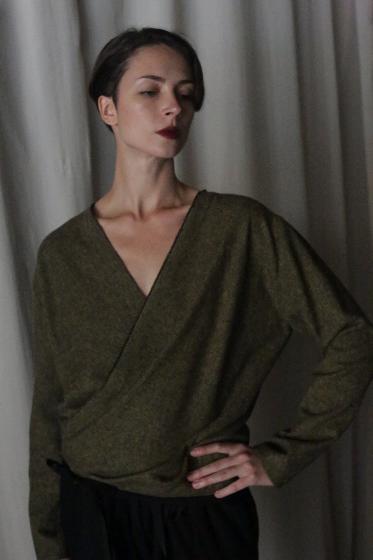A person with short dark hair stands with hand on hip, wearing the X Blouse With Sleeves in Brushed Cotton Moss Herringbone and dark pants. Light-colored curtains form the background. Their expression is thoughtful, with slightly closed eyes.