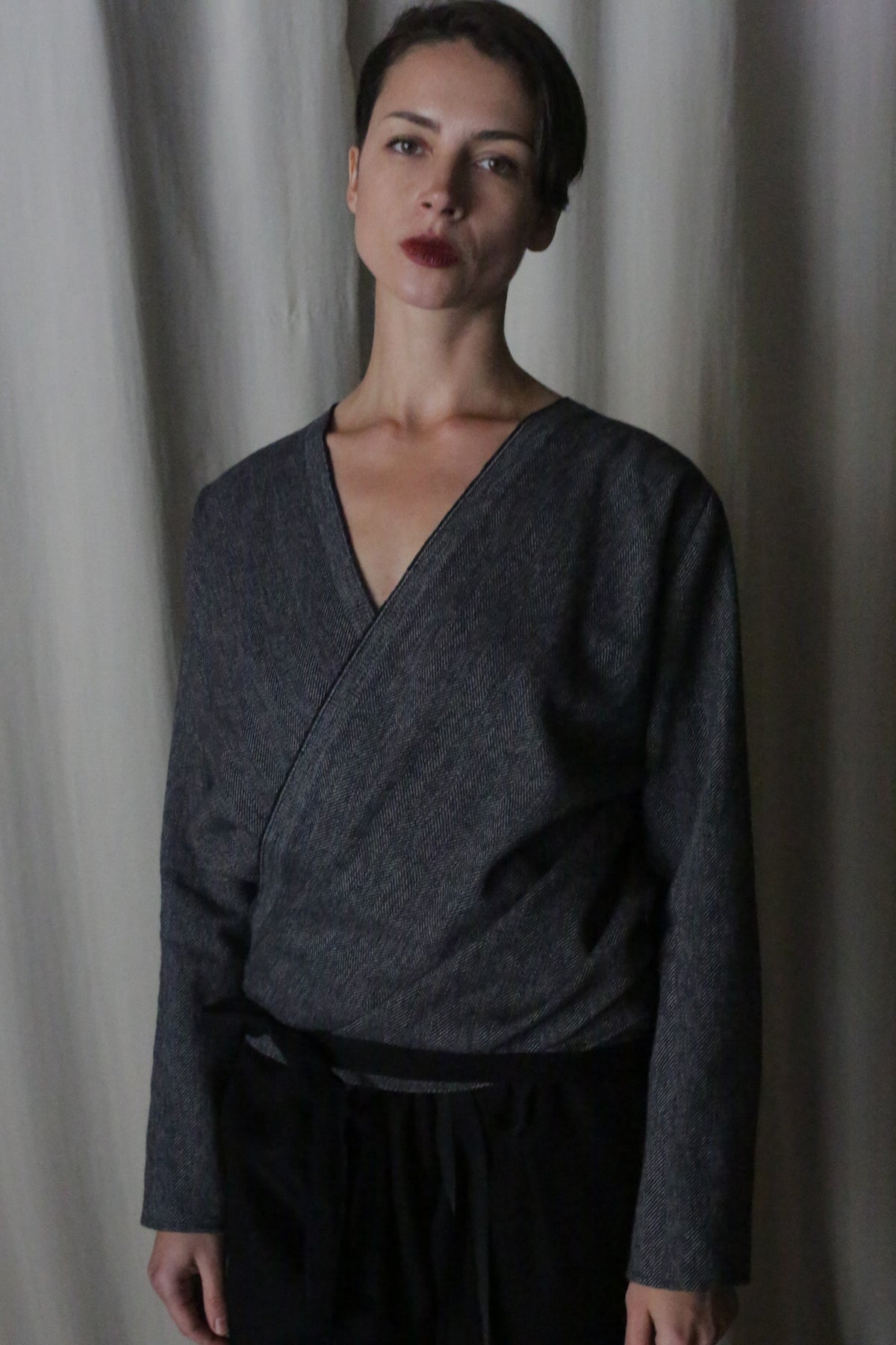 A person with short dark hair stands in front of a gray curtain, wearing a "X Blouse With Sleeves" made from brushed cotton in a blue hue, paired with black pants designed with low-waste principles, displaying a serious expression.