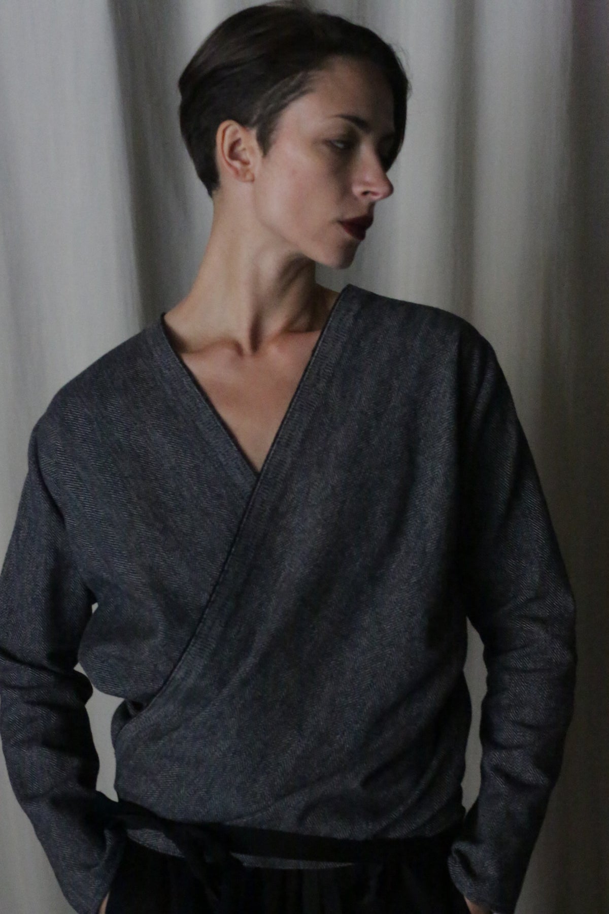 A person with short hair is wearing the X Blouse With Sleeves in Brushed Cotton Blue, standing against a neutral curtain backdrop. They are gazing to the side with a subtle, thoughtful expression.