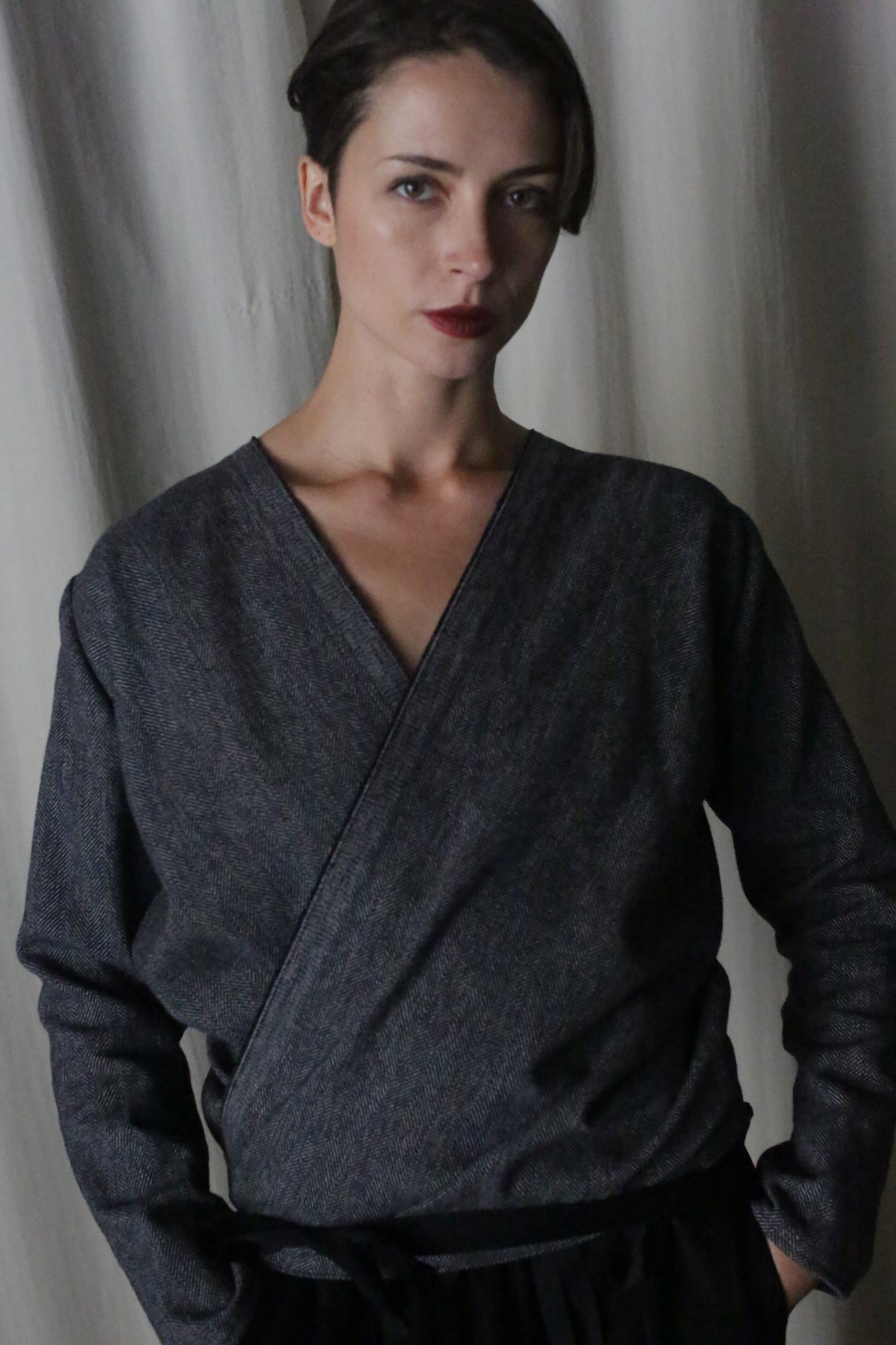 A woman with short brown hair wears a dark blue X Blouse With Sleeves in brushed cotton herringbone and stands in front of light-colored curtains, looking at the camera with a neutral expression.
