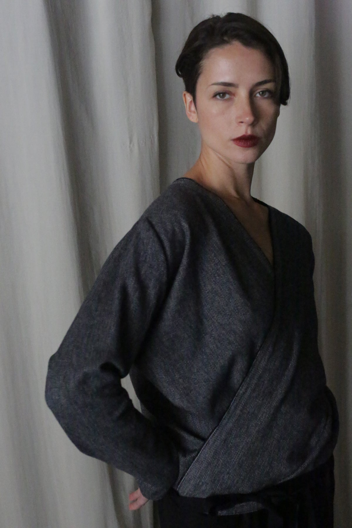 A woman with short dark hair and red lipstick stands before cream-colored curtains, confidently looking at the camera while wearing a handmade X Blouse With Sleeves in brushed cotton blue herringbone.