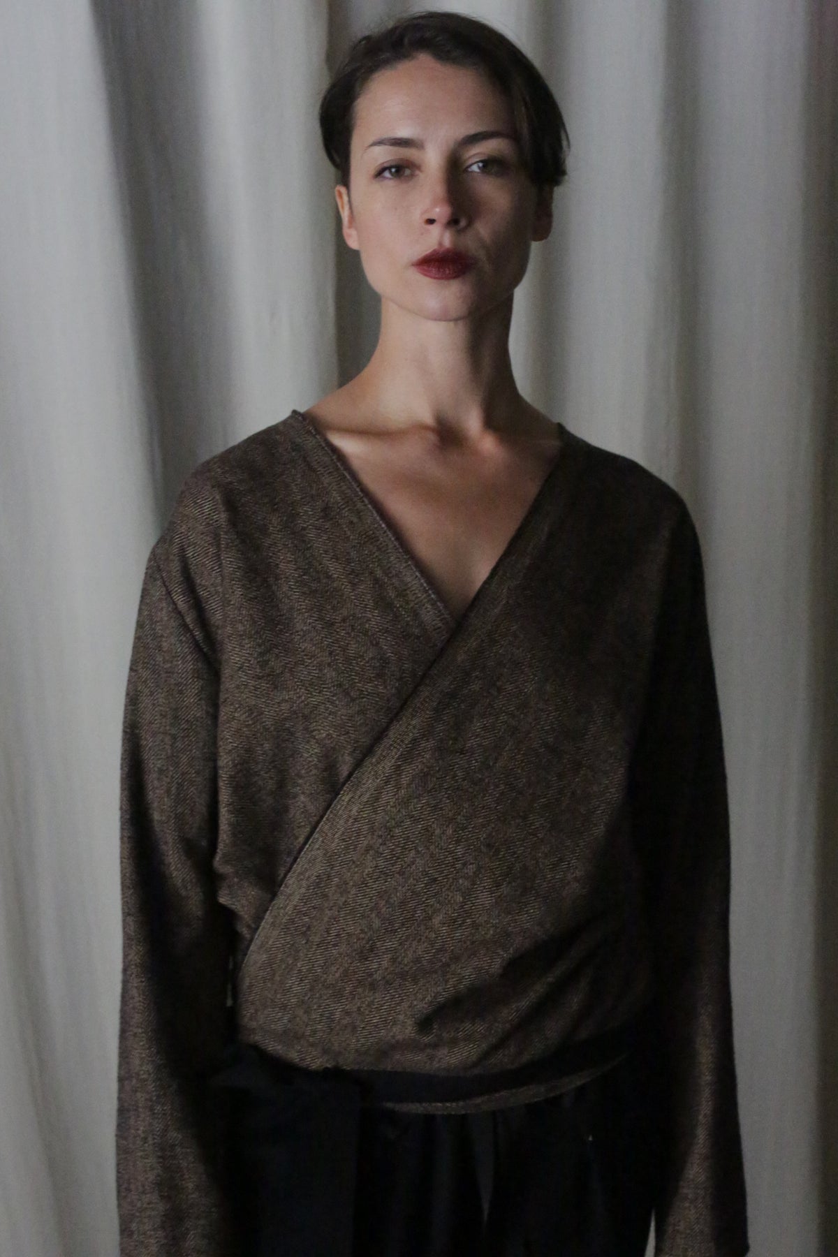 Standing against a neutral curtain backdrop is a person with short dark hair, wearing the X Blouse With Sleeves in Brushed Cotton Brown Herringbone. Their calm expression and maroon lipstick contrast the attire, highlighted by soft lighting that enhances this multifunctional wardrobe essential.
