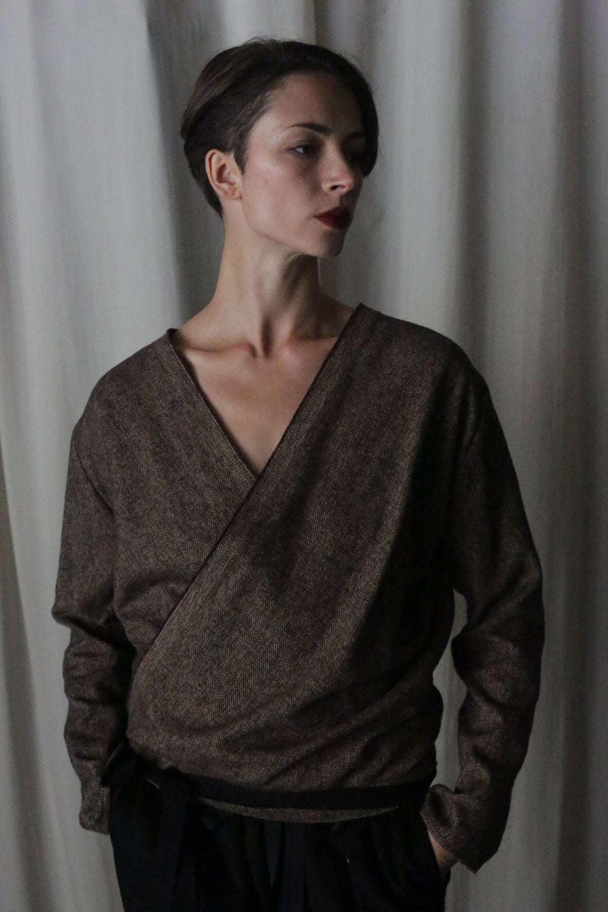 A person with short hair stands against a curtain backdrop wearing the X Blouse With Sleeves in brushed cotton brown herringbone, a multifunctional wardrobe essential featuring a deep V-neck. They have one hand in their pocket while gazing to the side.