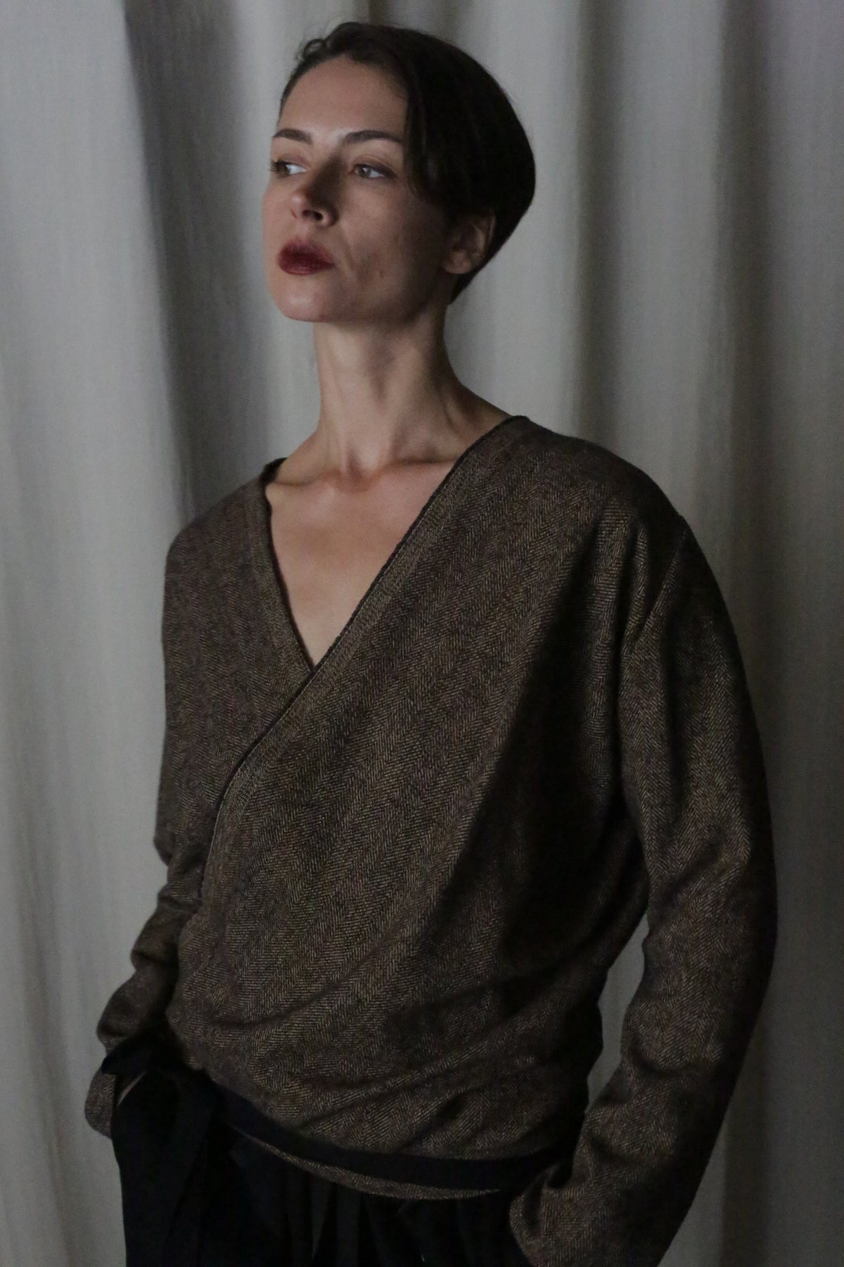 A person with short dark hair stands against a neutral curtain backdrop, wearing the X Blouse With Sleeves in brushed cotton brown herringbone and dark pants. This versatile piece enhances their contemplative expression as they tuck their hands into their pockets.