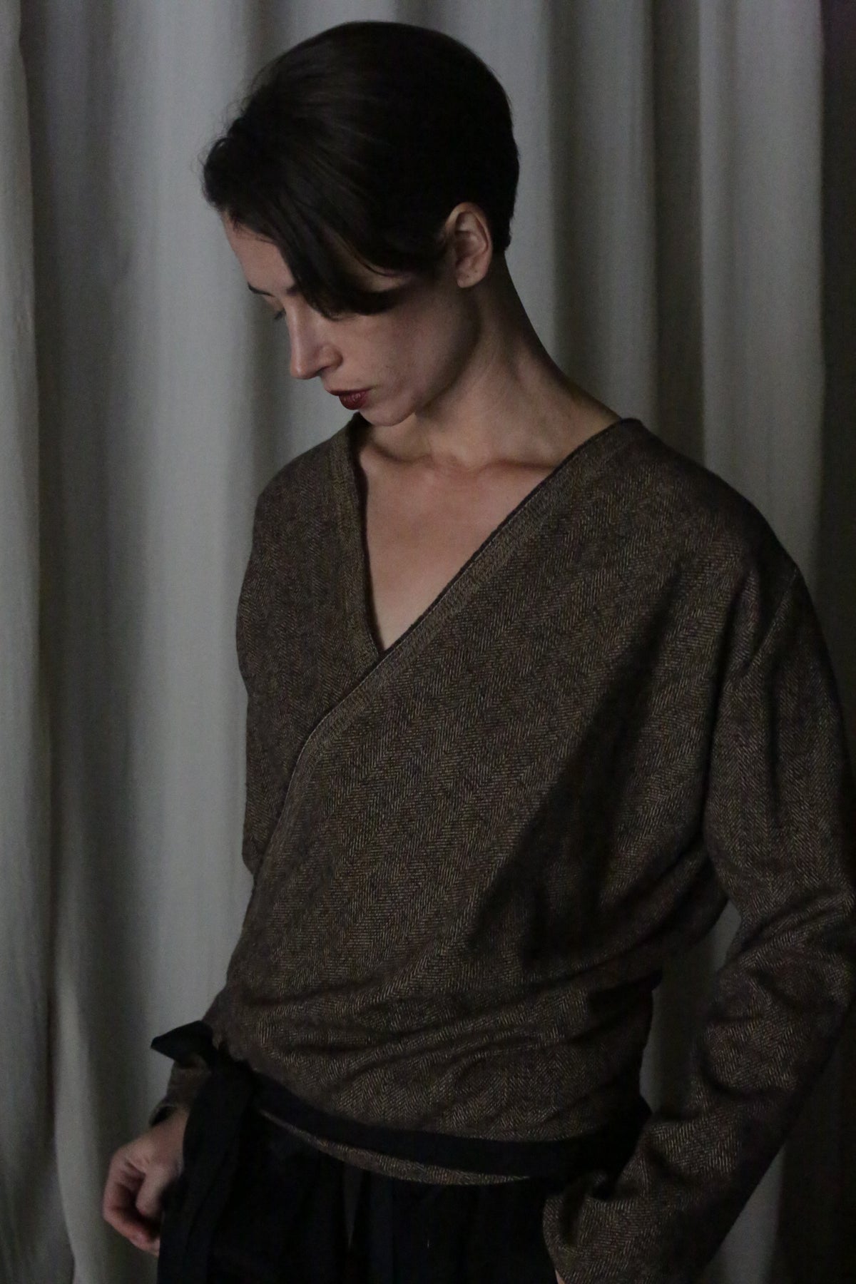 A person with short dark hair stands against draped white curtains, dressed in a multifunctional essential—the brushed cotton brown herringbone X Blouse With Sleeves. They look down thoughtfully, hands partially tucked in their pockets.