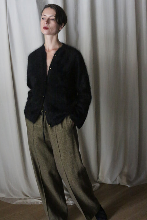 A person stands against a cream curtain, wearing a black fuzzy cardigan and high-waisted moss herringbone brushed cotton trousers. They have short hair, red lipstick, and pose with hands in pockets, gazing thoughtfully to the side, subtly nodding to Atelier Lan Jaenicke's elegant aesthetic.