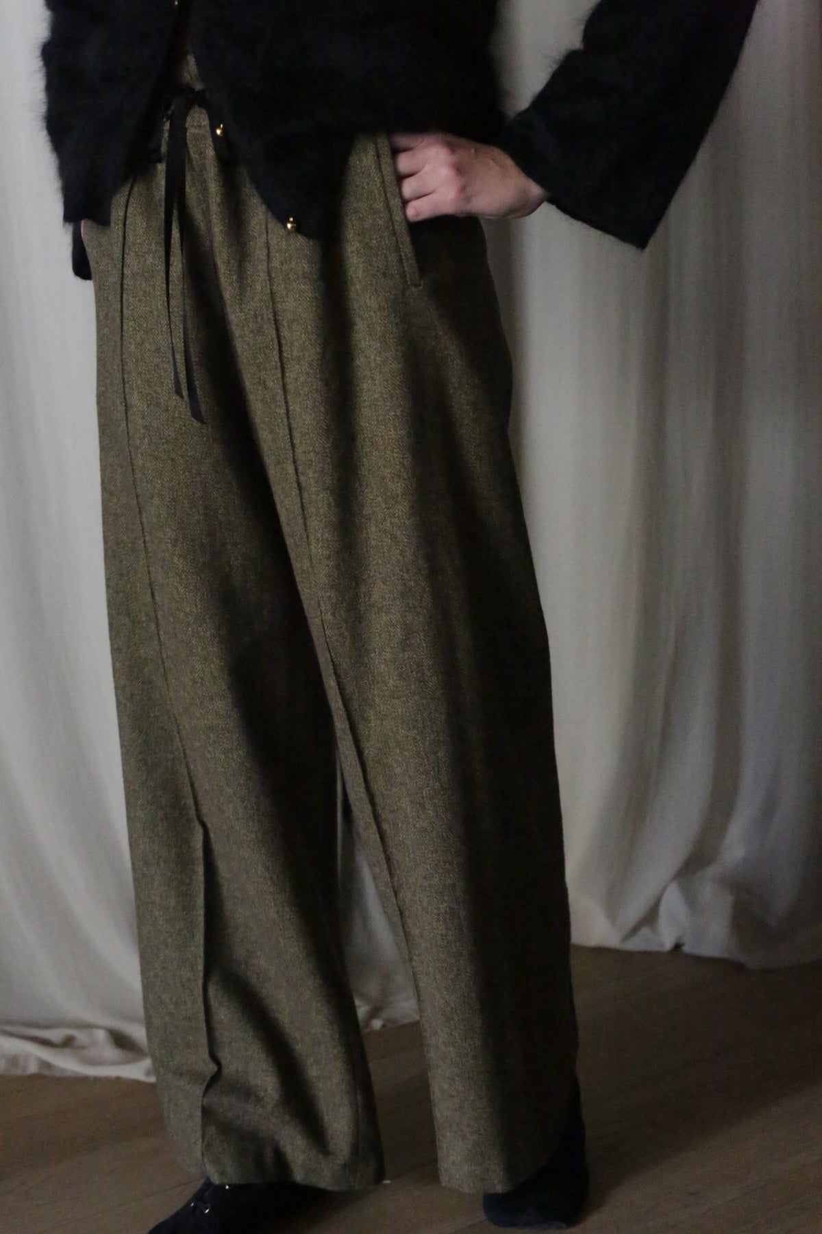 A person stands gracefully in front of a curtain, wearing elongating wide-legged Moss Herringbone trousers made from brushed cotton with hands in pockets and a black top.