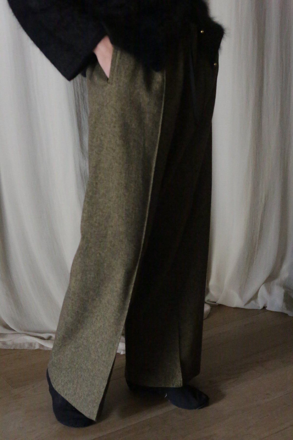 Wearing brushed cotton trousers in moss herringbone from Atelier Lan Jaenicke and a dark top, the person stands gracefully before white draped curtains on a wooden floor, with hands casually resting in pockets, complemented by dark socks.