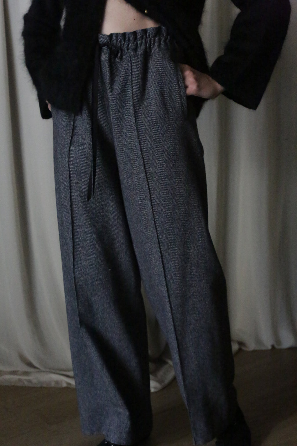 A person dons high-waisted, wide-legged gray pants made from brushed cotton twill with a black drawstring and a fuzzy black top. Their hands rest in the pockets against a light curtain backdrop, reminiscent of an Atelier Lan Jaenicke silhouette.