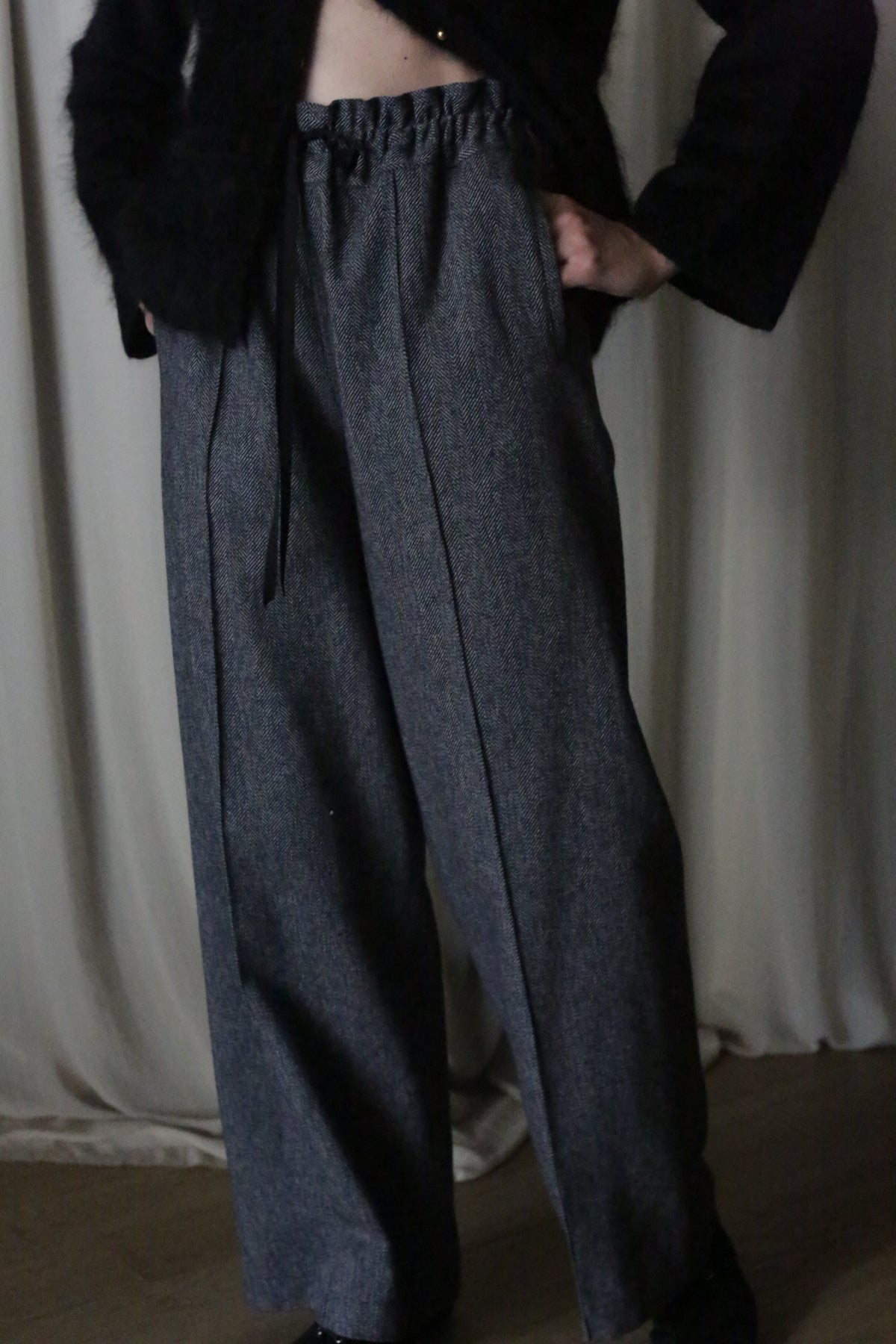 A person dons high-waisted, wide-legged gray pants made from brushed cotton twill with a black drawstring and a fuzzy black top. Their hands rest in the pockets against a light curtain backdrop, reminiscent of an Atelier Lan Jaenicke silhouette.
