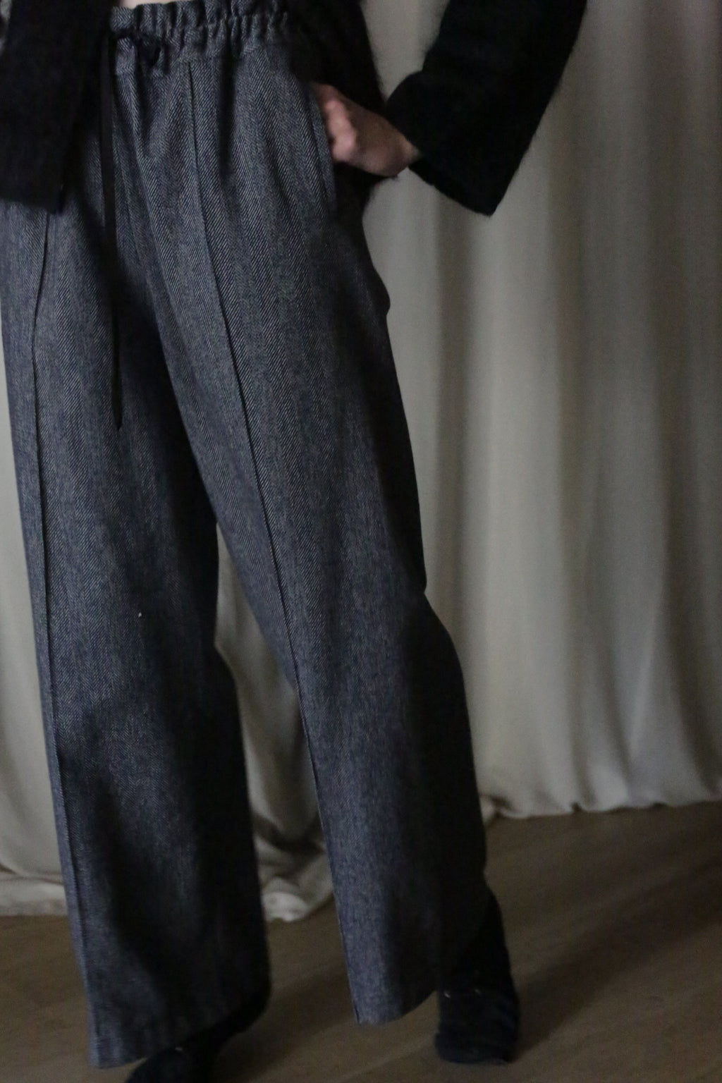 Against a cream curtain backdrop, a person on a wooden floor wears blue herringbone straight-leg trousers with an elastic waistband from Atelier Lan Jaenicke. With hands in pockets and paired with a dark top and black shoes, the dim lighting enhances the ensemble's elegance.