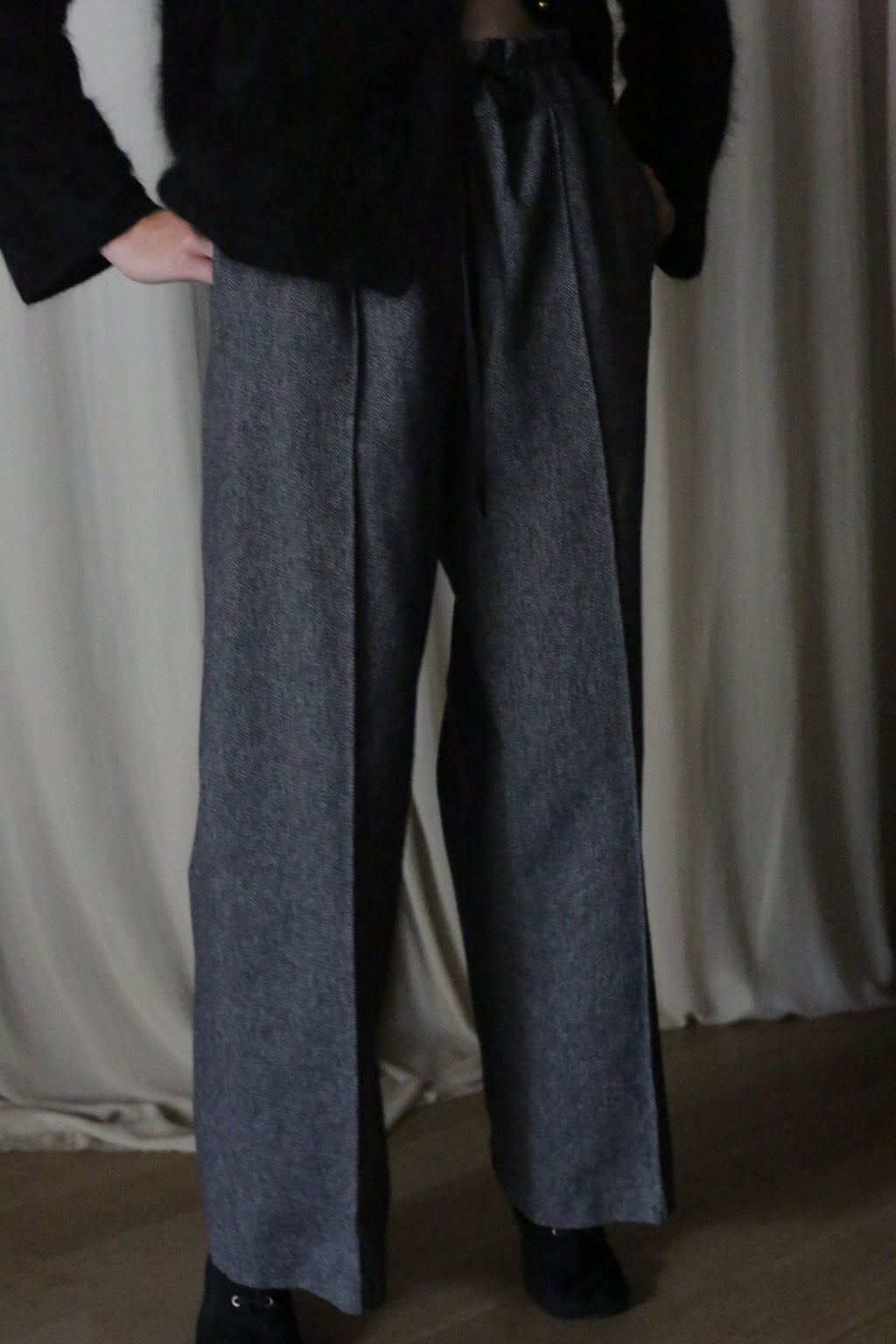 Wearing Brushed Cotton Trousers in Blue Herringbone, a person with a dark top stands against curtains. Hands in pockets, dim lighting casts shadows.