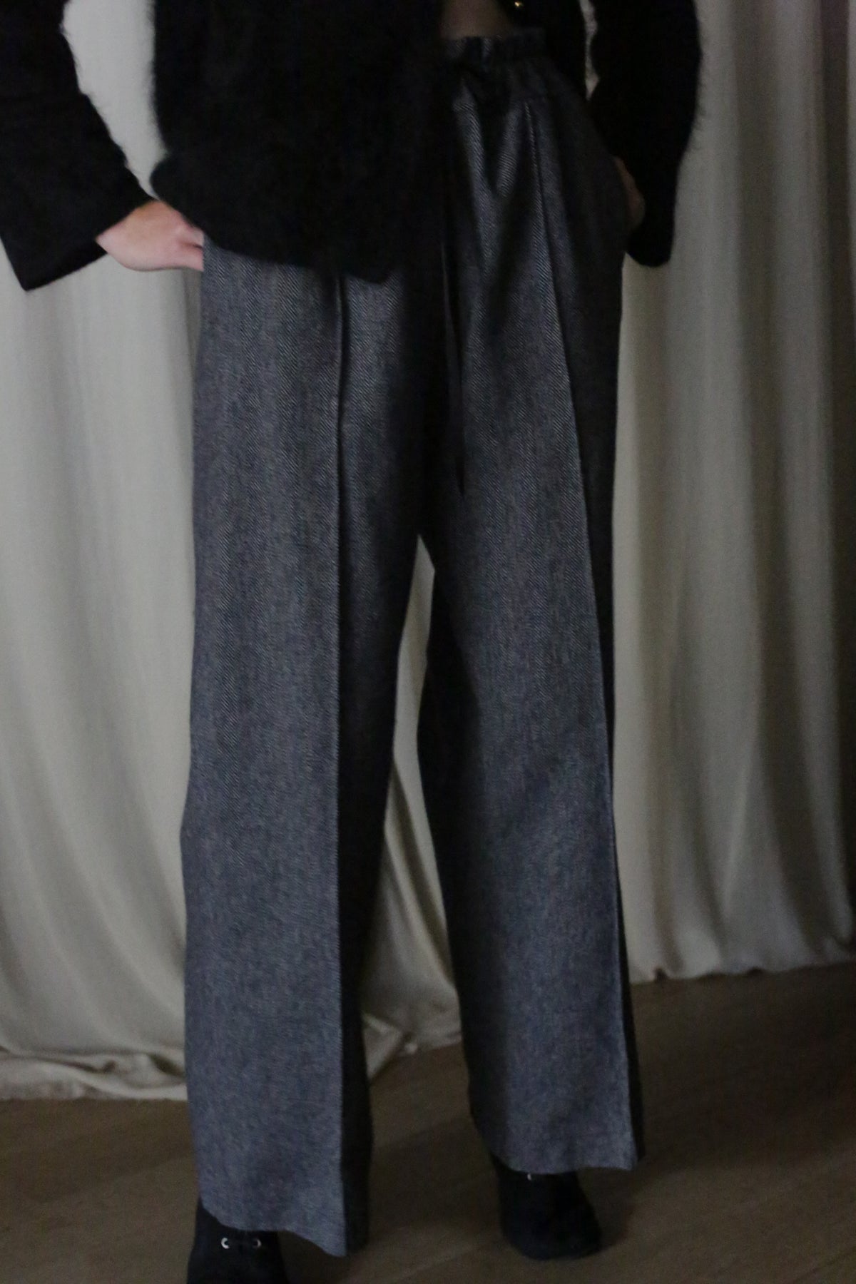 Wearing Brushed Cotton Trousers in Blue Herringbone, a person with a dark top stands against curtains. Hands in pockets, dim lighting casts shadows.