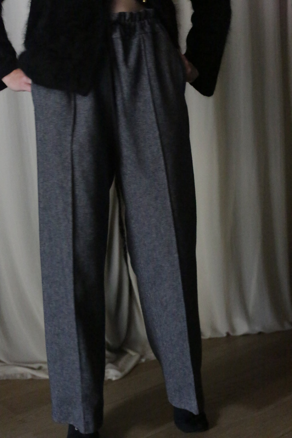 A person stands in Atelier Lan Jaenicke's Brushed Cotton Trousers in Blue Herringbone, hands casually tucked in pockets. The cotton twill pants complement their dark top, while a light curtain gracefully frames the wooden floor.