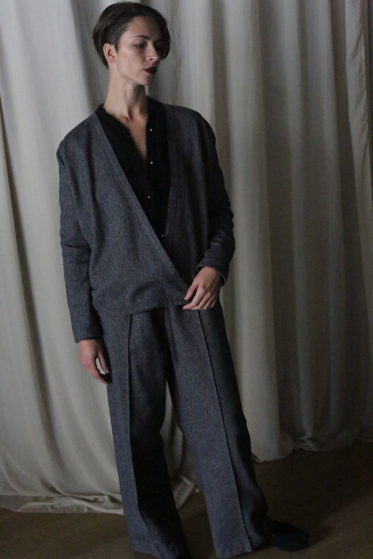 A person with short hair stands before cream curtains, wearing a dark gray pantsuit over a black top. Their left arm rests by their side, showcasing the elongating straight-leg design of the Atelier Lan Jaenicke suit.