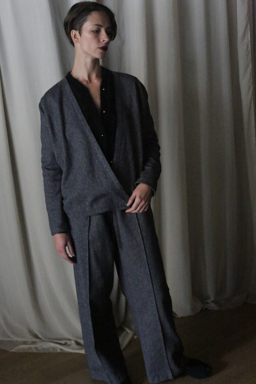 A person with short hair stands before cream curtains, wearing a dark gray pantsuit over a black top. Their left arm rests by their side, showcasing the elongating straight-leg design of the Atelier Lan Jaenicke suit.