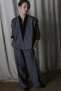 A person stands against a dimly lit curtain in an Atelier Lan Jaenicke suit, featuring a loose-fitting blazer and Brushed Cotton Trousers in blue herringbone. Their hands are in pockets, gazing downward with neatly styled short hair, creating a soft, moody ambiance.