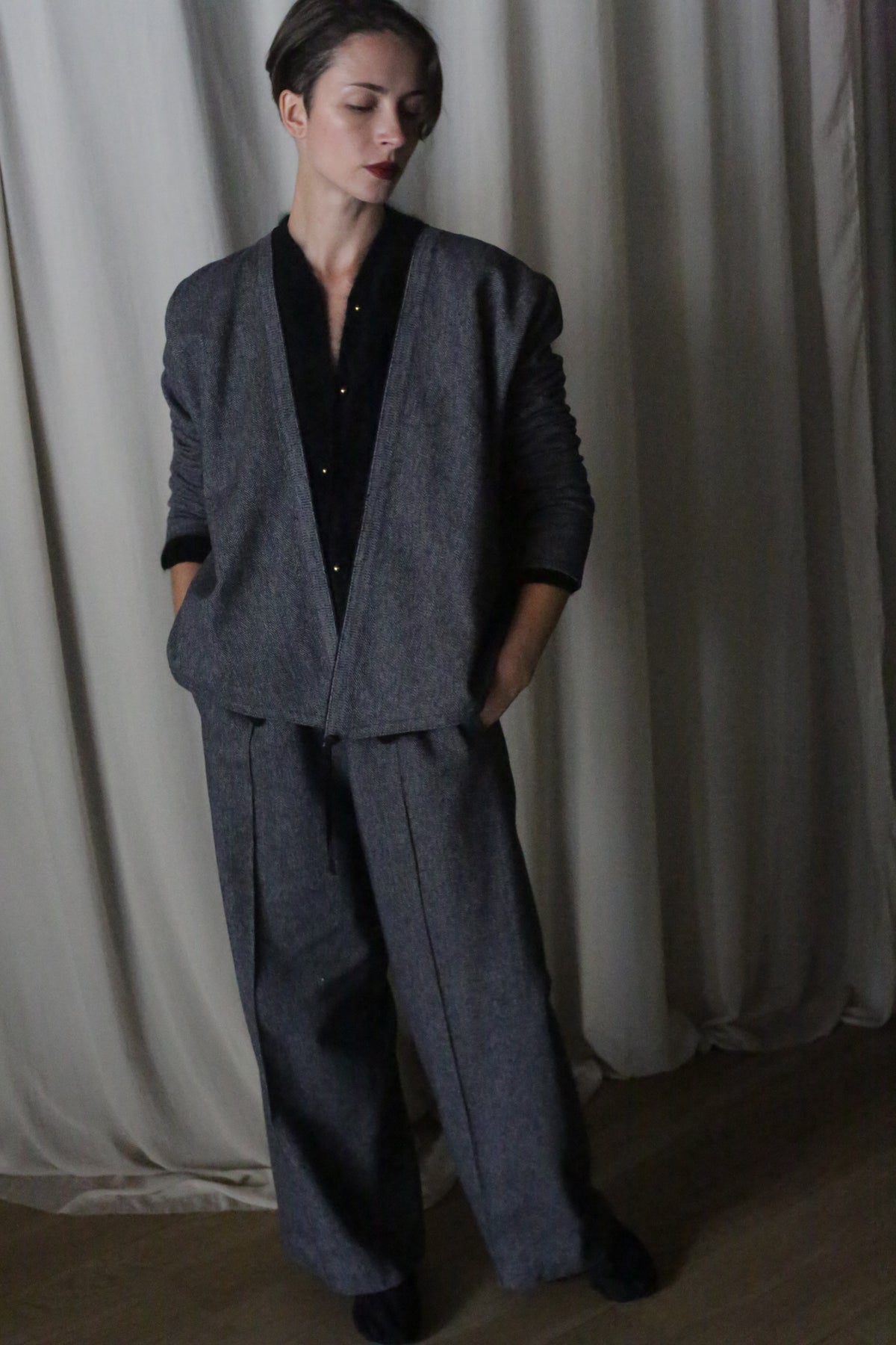 A person stands indoors by light curtains, wearing a dark gray suit paired with the X Blouse With Sleeves in brushed cotton blue herringbone. Hands in pockets, short-haired, they look downward with a calm expression.