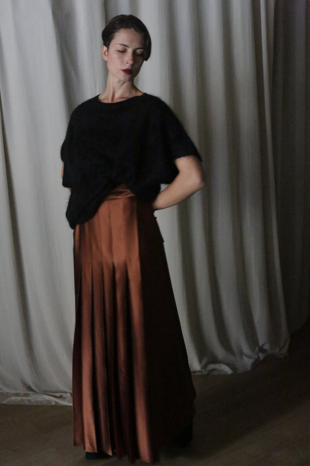 A woman stands against draped curtains, wearing a black top and the handmade Copper Pleated Layered Skirt that flows elegantly. Her hair is styled up, and she gazes slightly downwards with a calm expression.