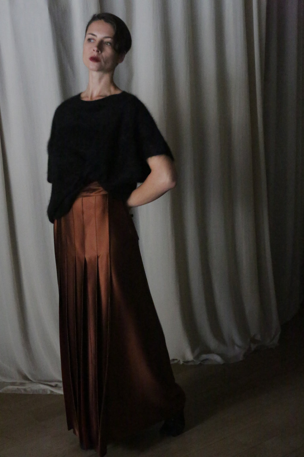 A woman stands gracefully in front of a neutral curtain, wearing a dark short-sleeve top paired with the Copper Pleated Layered Skirt from Atelier Lan Jaenicke. Her hand rests on her hip in an indoor setting that captures the sophisticated artistry of San Francisco.