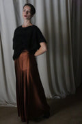 A woman stands gracefully in front of a neutral curtain, wearing a dark short-sleeve top paired with the Copper Pleated Layered Skirt from Atelier Lan Jaenicke. Her hand rests on her hip in an indoor setting that captures the sophisticated artistry of San Francisco.