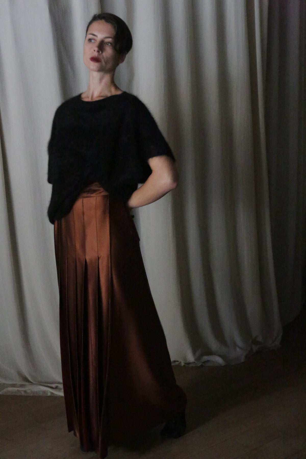 A woman stands gracefully in front of a neutral curtain, wearing a dark short-sleeve top paired with the Copper Pleated Layered Skirt from Atelier Lan Jaenicke. Her hand rests on her hip in an indoor setting that captures the sophisticated artistry of San Francisco.
