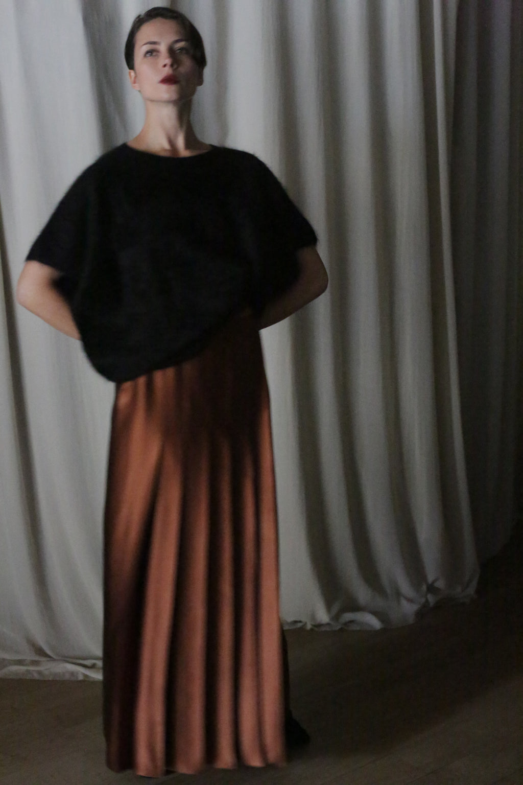 A person stands indoors against a backdrop of draped silk charmeuse fabric. They wear a black top and the Copper Pleated Layered Skirt, a long, flowing brown skirt from Atelier Lan Jaenicke in San Francisco. With their hands on their hips, they gaze upwards.