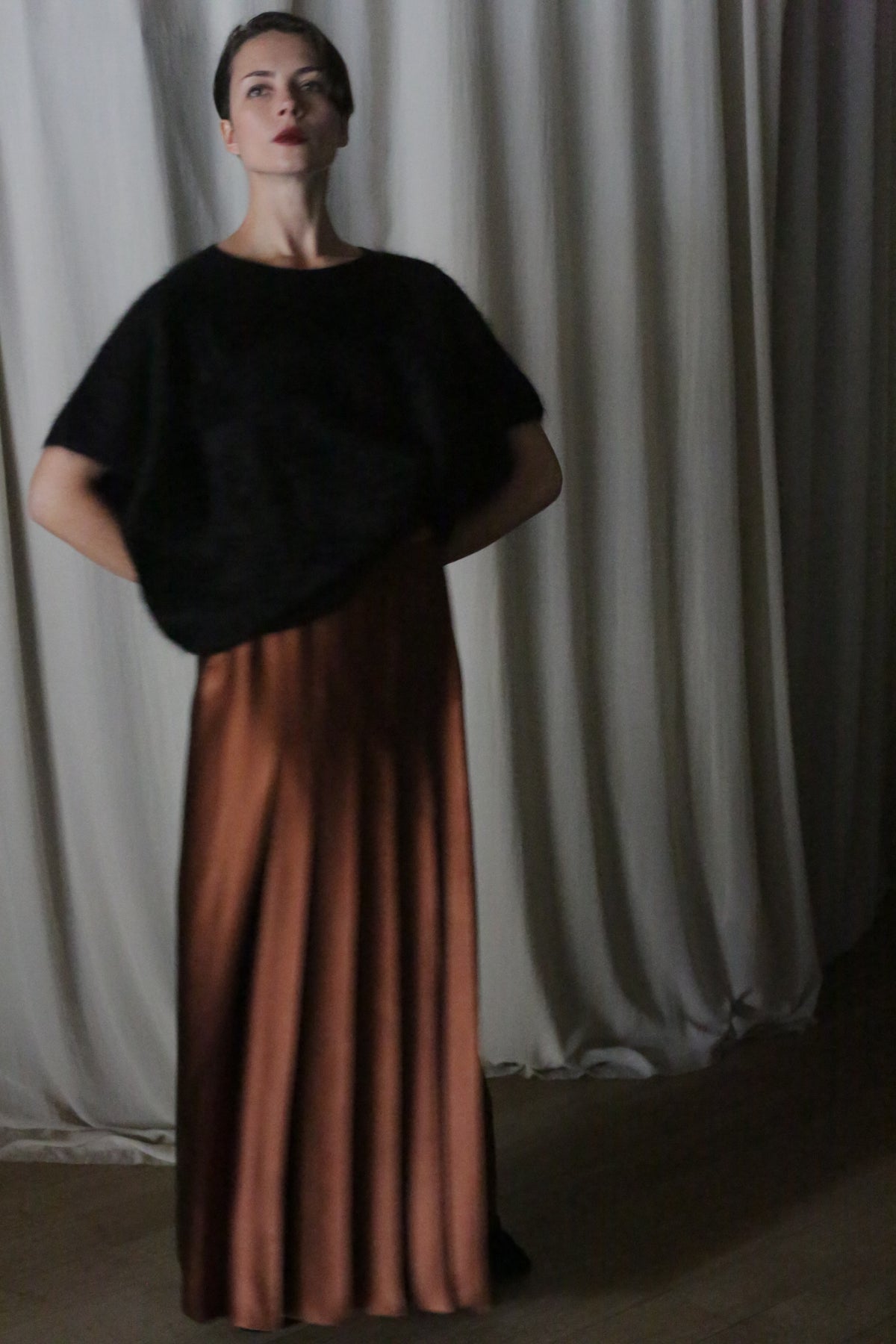 A person stands indoors against a backdrop of draped silk charmeuse fabric. They wear a black top and the Copper Pleated Layered Skirt, a long, flowing brown skirt from Atelier Lan Jaenicke in San Francisco. With their hands on their hips, they gaze upwards.