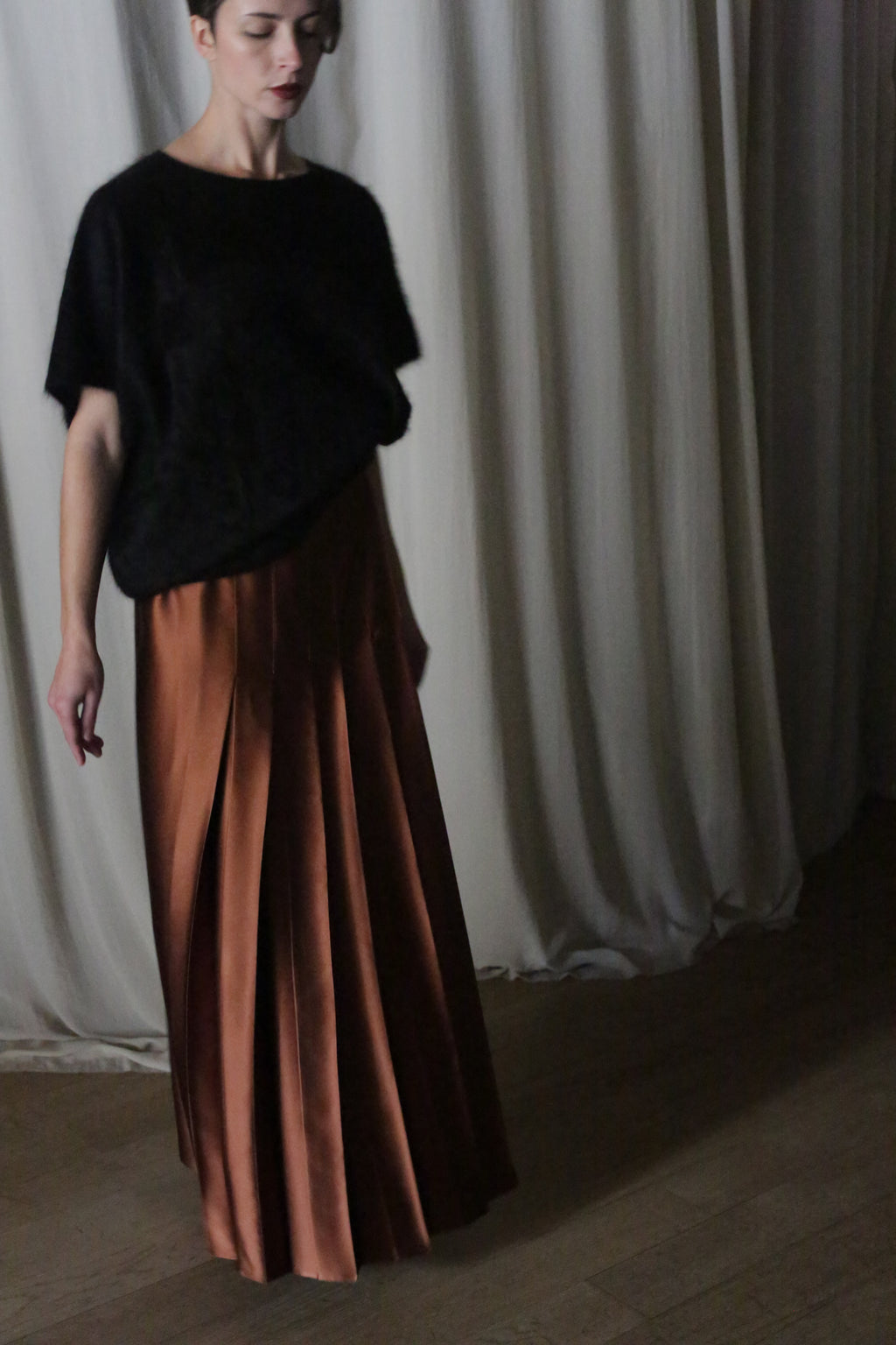 A woman wearing a black top and a Copper Pleated Layered Skirt, crafted from heavy silk charmeuse, stands gracefully before neutral drapes.