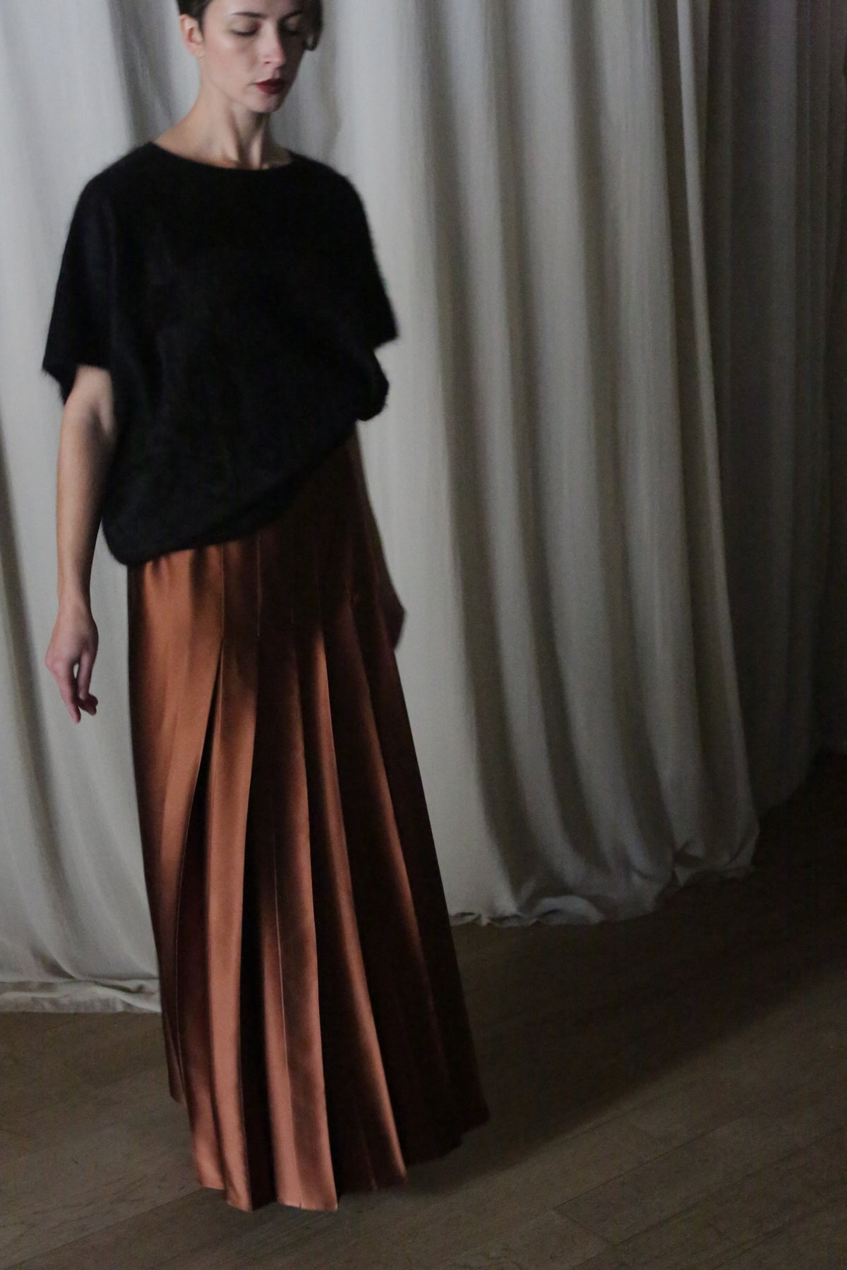 A woman wearing a black top and a Copper Pleated Layered Skirt, crafted from heavy silk charmeuse, stands gracefully before neutral drapes.