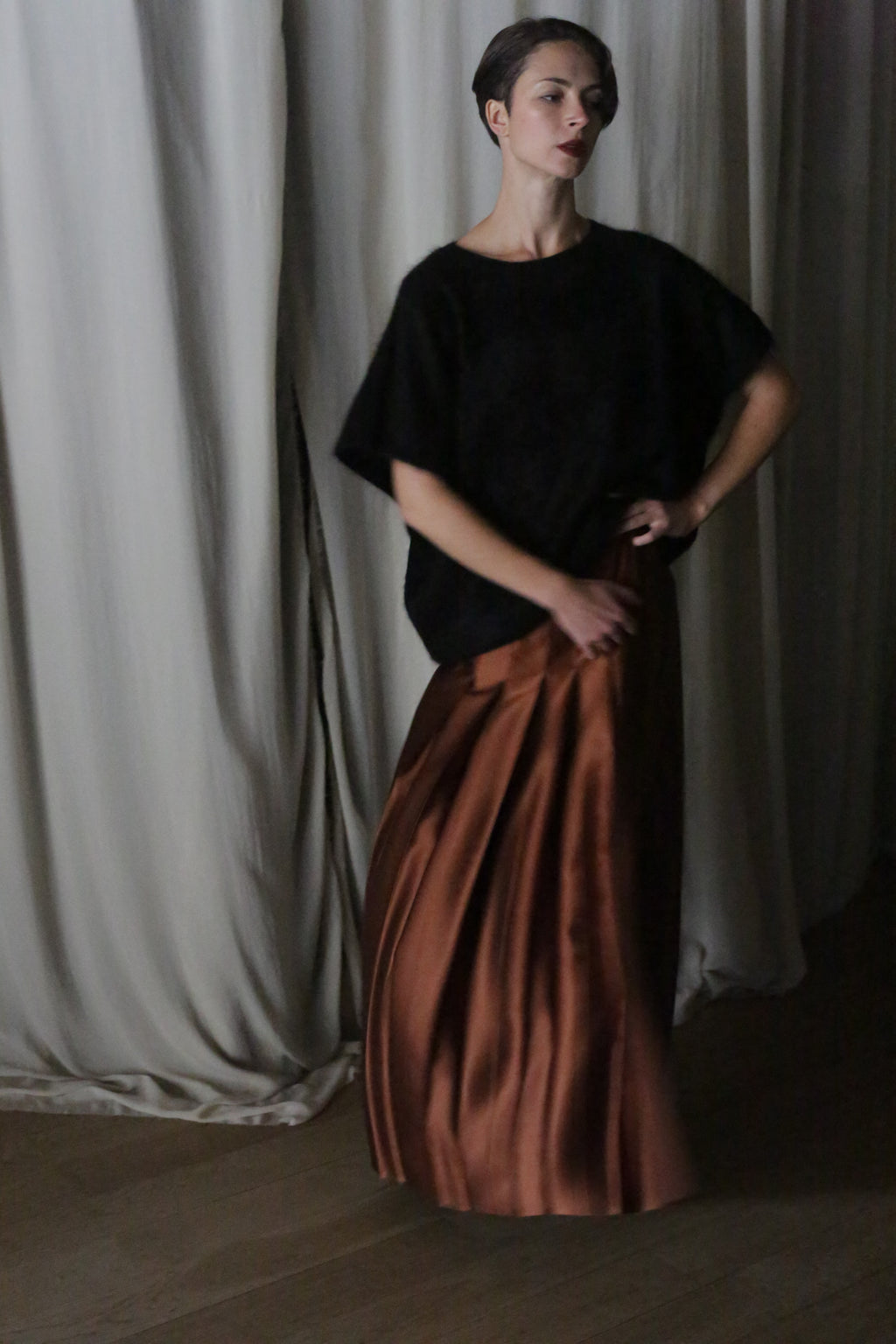 A person stands against draped white curtains, wearing a dark loose top and a handmade Copper Pleated Layered Skirt. With one hand on their hip, the pose, combined with the dim lighting, creates a moody atmosphere.