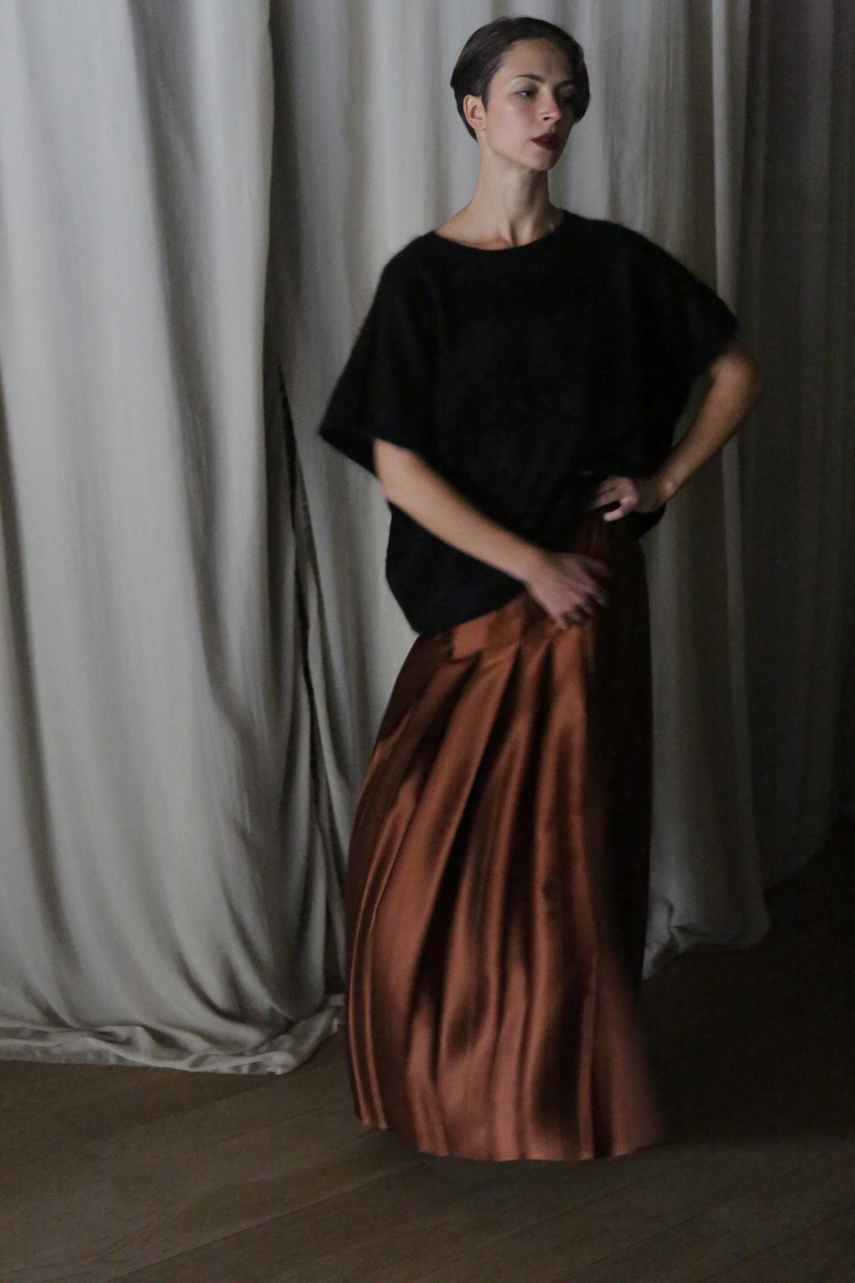 A person stands against draped white curtains, wearing a dark loose top and a handmade Copper Pleated Layered Skirt. With one hand on their hip, the pose, combined with the dim lighting, creates a moody atmosphere.
