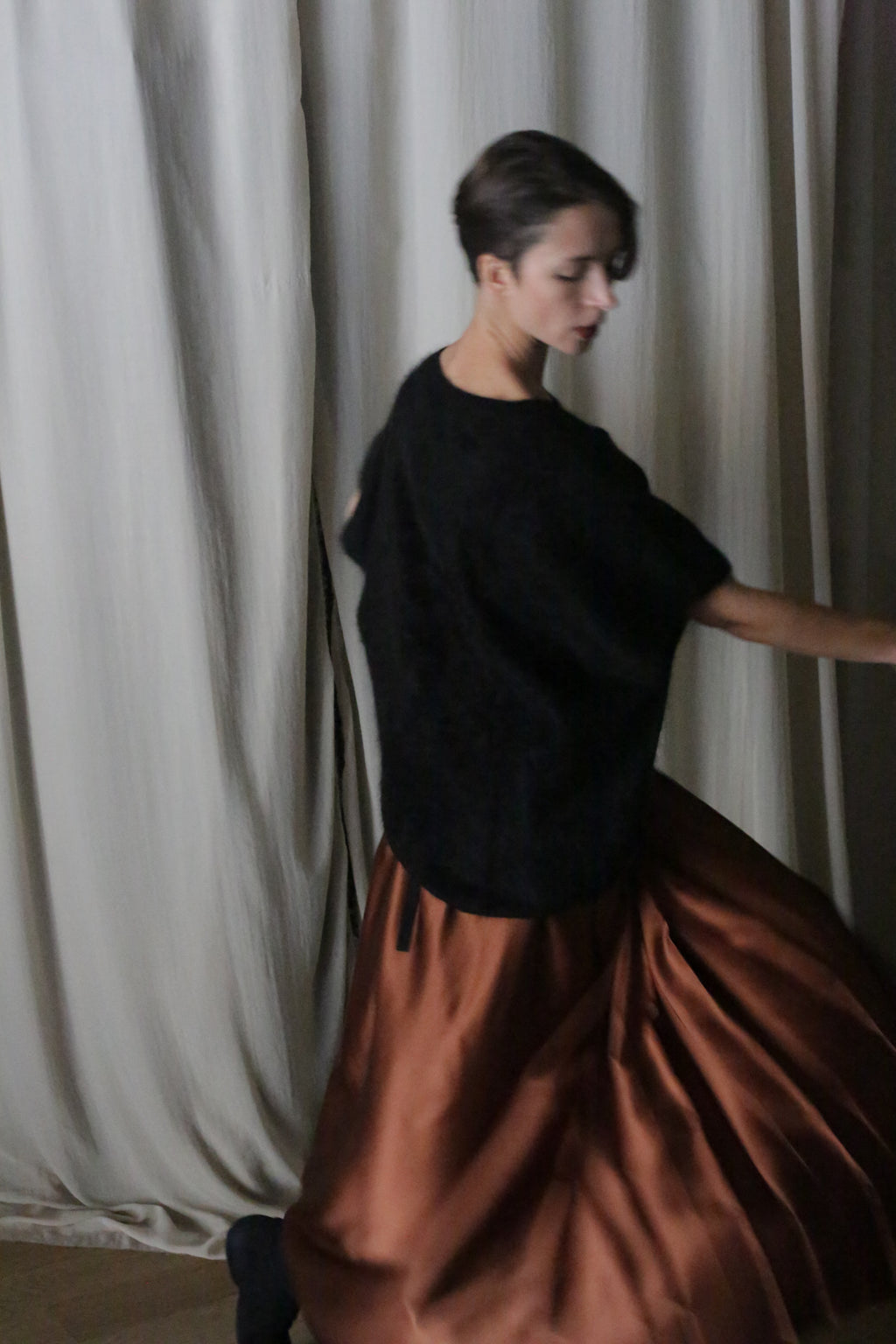A person spins gracefully in front of cream curtains, wearing a black top and a Copper Pleated Layered Skirt crafted from heavy silk charmeuse. Their short hair and the skirt's handmade in-house design enhance the dynamic movement captured in the image.