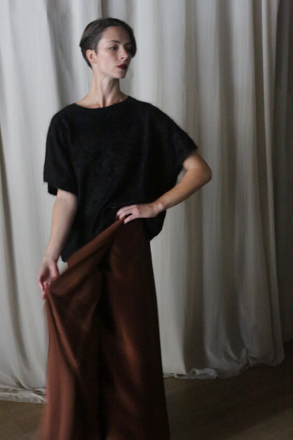 Against a backdrop of white curtains, a person wearing a black top holds the hem of their handmade Copper Pleated Layered Skirt. With short hair and a poised side glance, they stand in soft lighting that enhances the serene atmosphere.