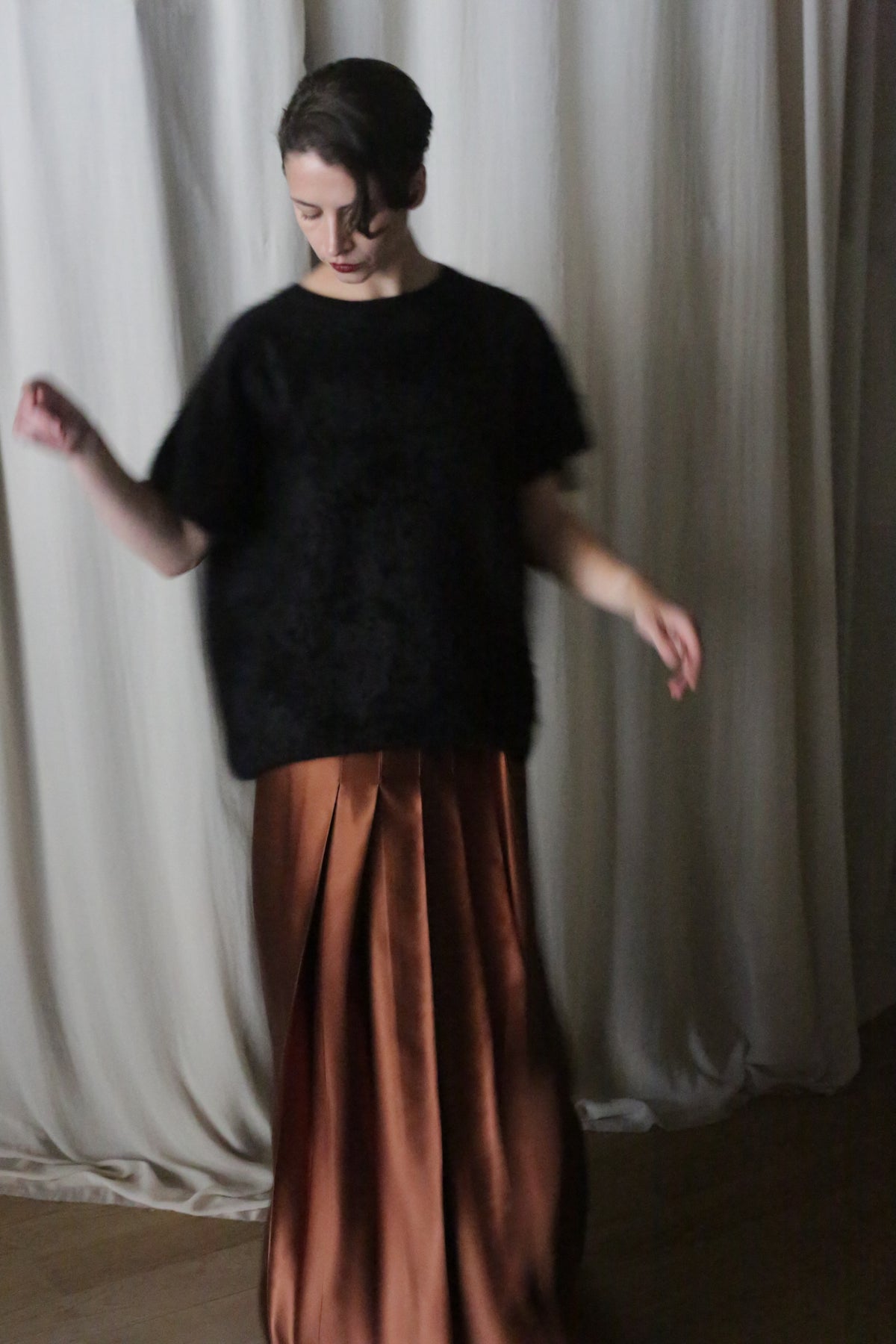A person stands in front of white curtains, wearing a black top and a flowing Atelier Lan Jaenicke Copper Pleated Layered Skirt. Their head is slightly turned and their arms are minimally extended, suggesting a gentle movement or dance. The silk charmeuse fabric catches the soft San Francisco lighting beautifully.