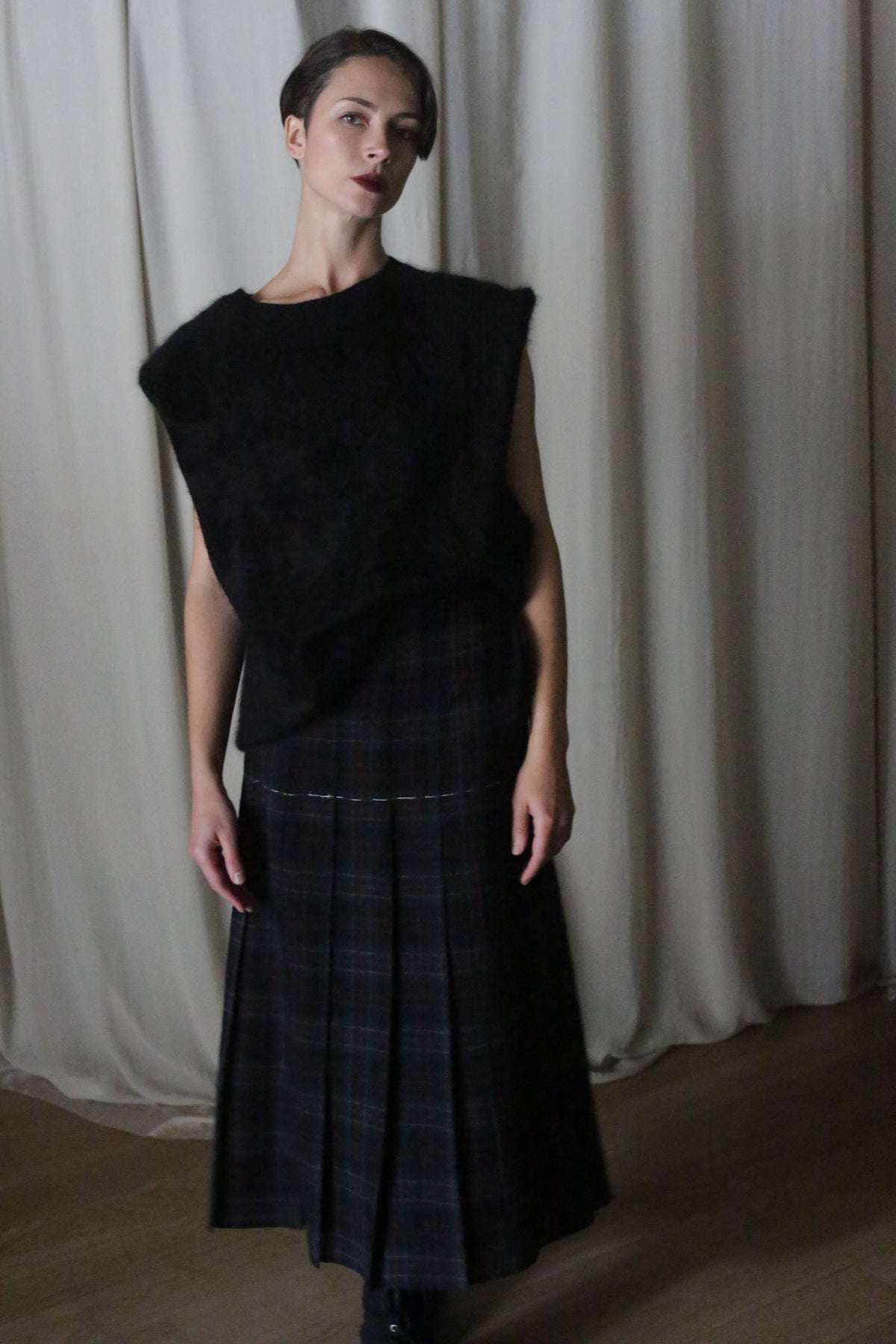 A person with short hair stands indoors in moody, dim lighting, wearing a sleeveless black textured top and a Hand Pleated Wrap Skirt in Japanese Wool Gauze Forest Plaid, set against light-colored curtains.