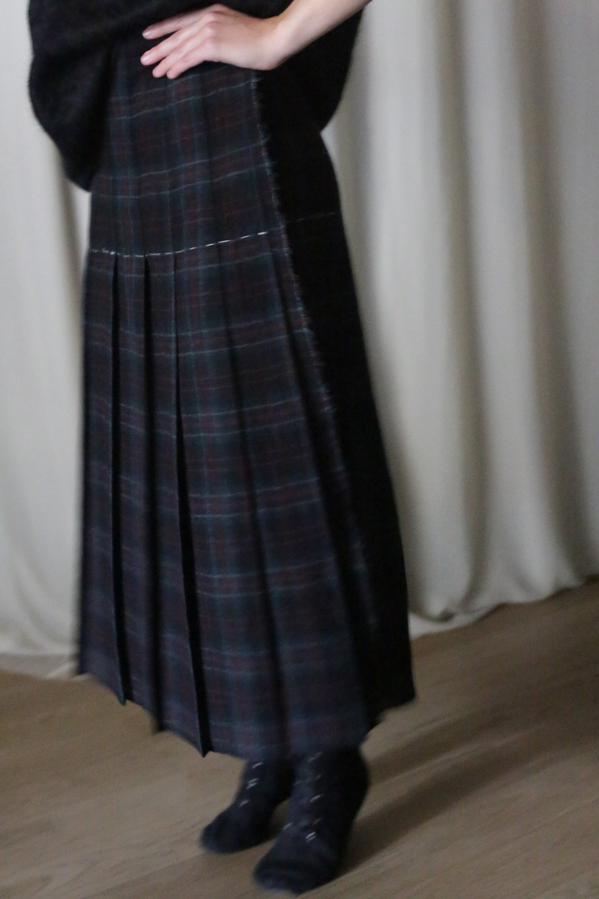 A person wearing the Hand Pleated Wrap Skirt | Japanese Wool Gauze Forest Plaid and black shoes stands with one hand on their hip before light curtains, with only their lower body visible.