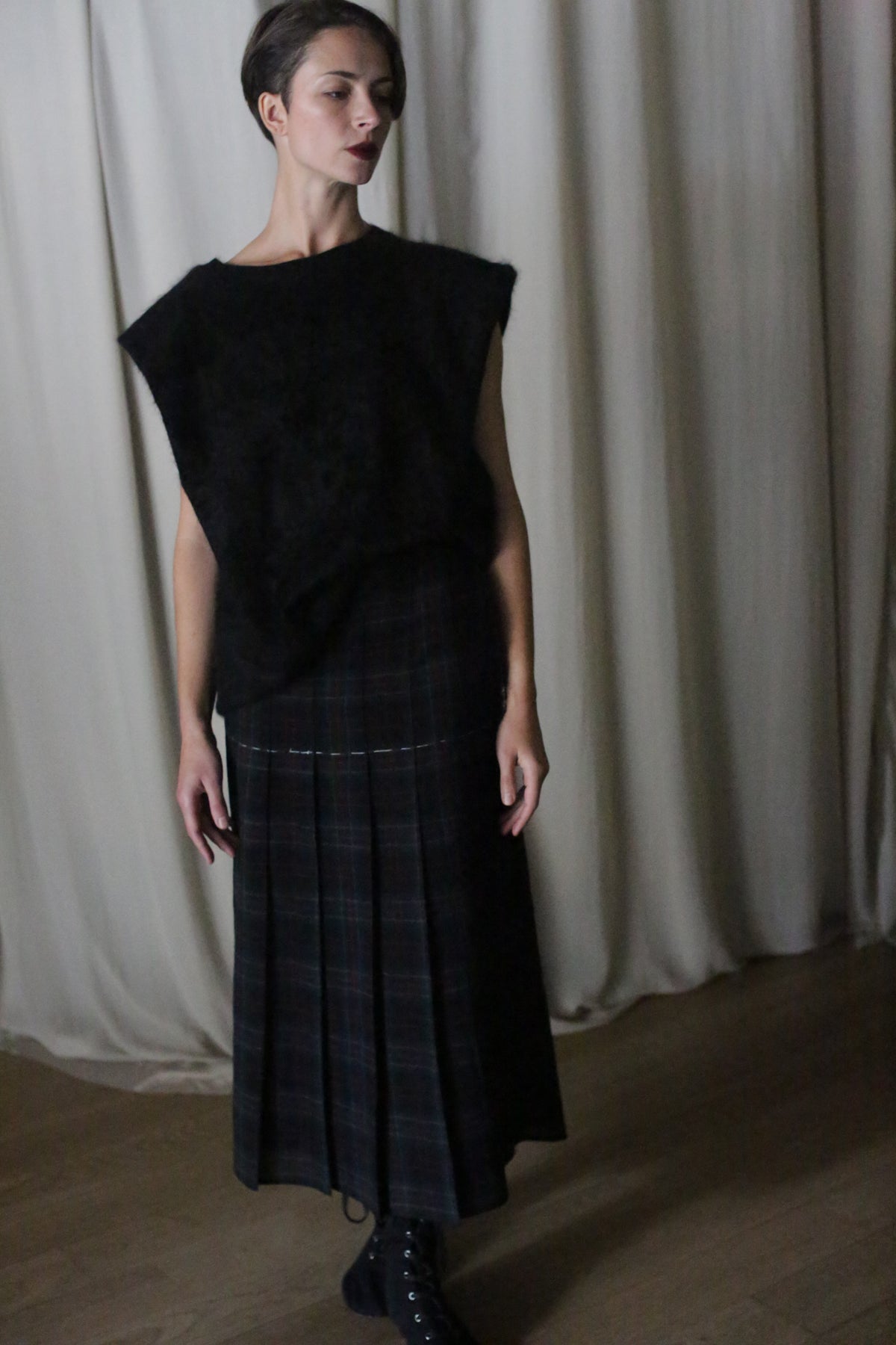 A person indoors wears a sleeveless black top, the Hand Pleated Wrap Skirt in Japanese Wool Gauze Forest Plaid, and black boots. They have short hair and dark lipstick, standing before light curtains on a wooden floor.