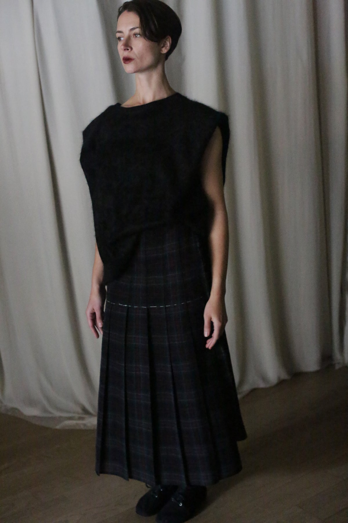 A woman with short, swept-back hair stands indoors before beige curtains in a dark sleeveless top and the Hand Pleated Wrap Skirt | Japanese Wool Gauze Forest Plaid, highlighting its refined, zero-waste design.