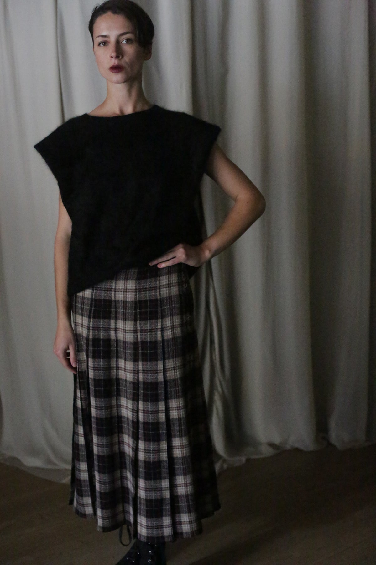 Standing confidently indoors with a hand on their hip, the person is dressed in a black top paired with a Hand Pleated Wrap Skirt made from Japanese Wool Gauze, showcasing a Taupe Plaid pattern. The scene is set against light-colored curtains in the background.