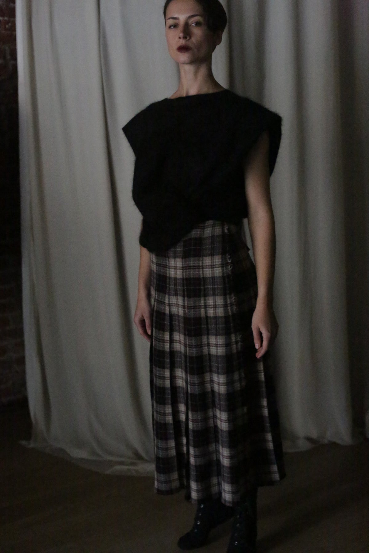 In a dimly lit room, an individual wears a handmade black top paired with the Hand Pleated Wrap Skirt crafted from Japanese Wool Gauze Plaid in taupe. The background reveals a curtain and part of a brick wall.
