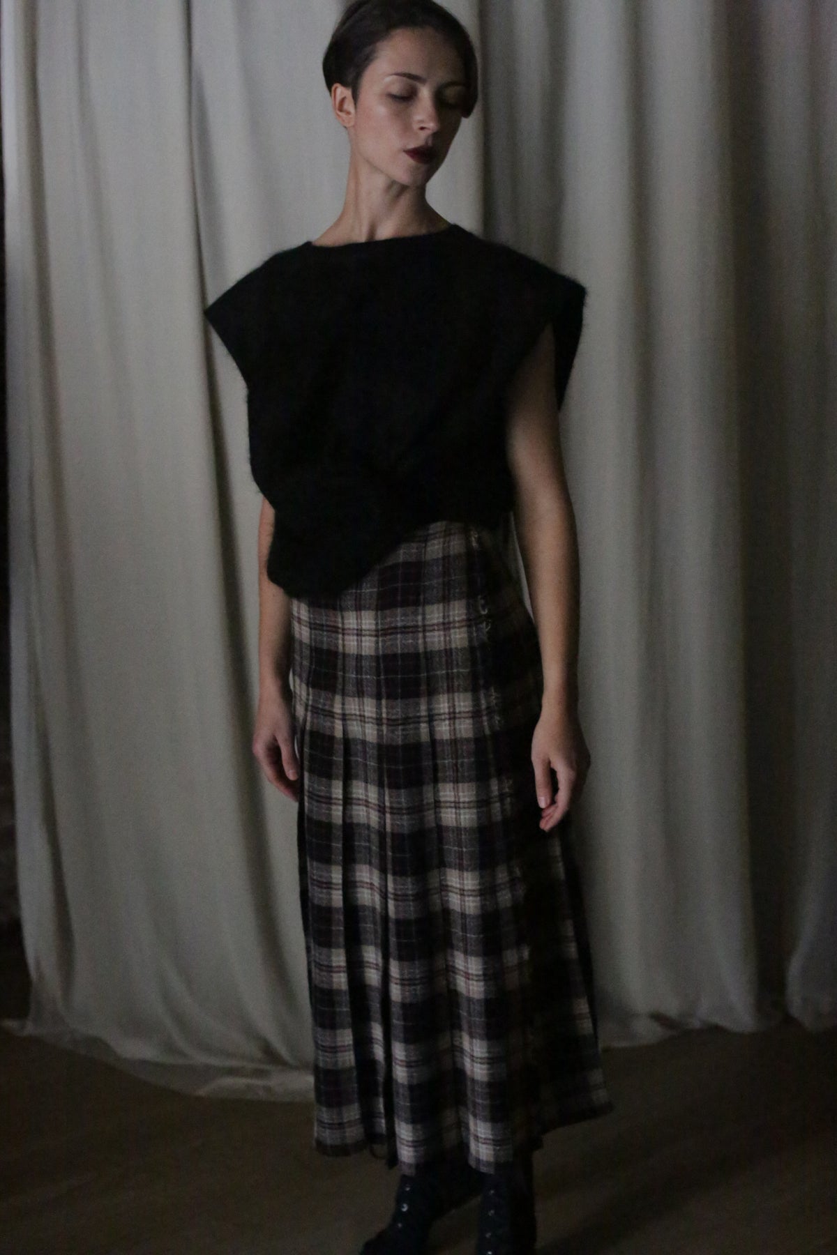 A person stands against a light curtain background wearing a sleeveless black top and the Hand Pleated Wrap Skirt made from Japanese Wool Gauze Plaid Taupe. They have short dark hair and are looking downward, with dim lighting enhancing the moody atmosphere, highlighting the beauty of zero-waste design.