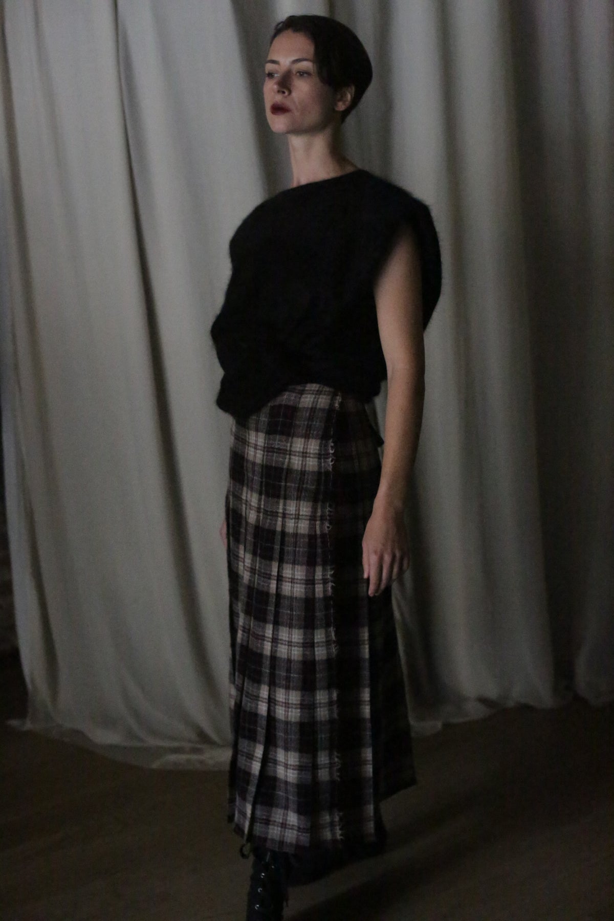 A person with short hair stands before a curtain in dim light, wearing a sleeveless black top and the Hand Pleated Wrap Skirt in Japanese Wool Gauze Taupe Plaid, highlighting its zero-waste design.
