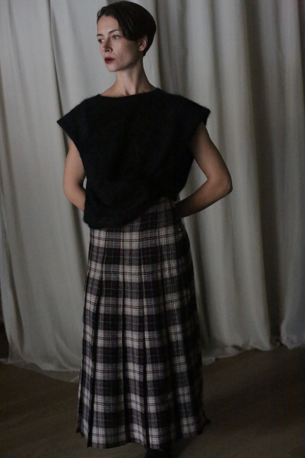 A person stands indoors against a curtain backdrop, wearing a black top and a long Hand Pleated Wrap Skirt made from Japanese Wool Gauze Taupe Plaid. Their hands are behind their back, creating intrigue in the dimly lit scene where soft shadows gently dance around them.