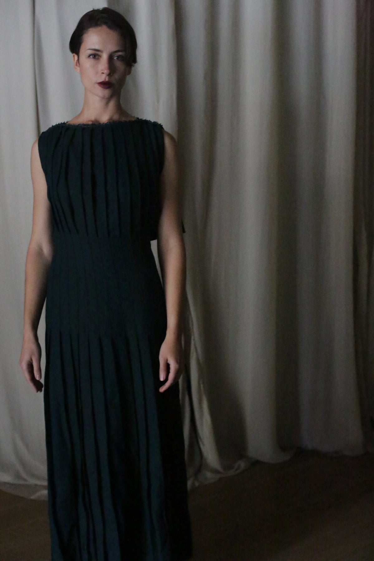 A woman with dark hair in an updo wears the Hand Pleated Column Dress in Japanese Wool Gauze Forest, a long, sleeveless gown with elegant pleats, standing before beige curtains and gazing forward neutrally.