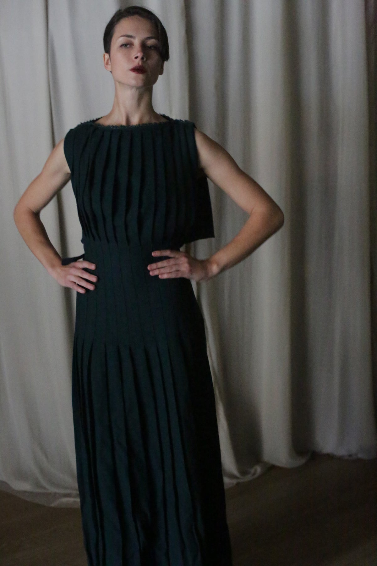 A woman stands confidently with hands on hips against light-colored curtains, wearing the Hand Pleated Column Dress in Japanese Wool Gauze Forest.