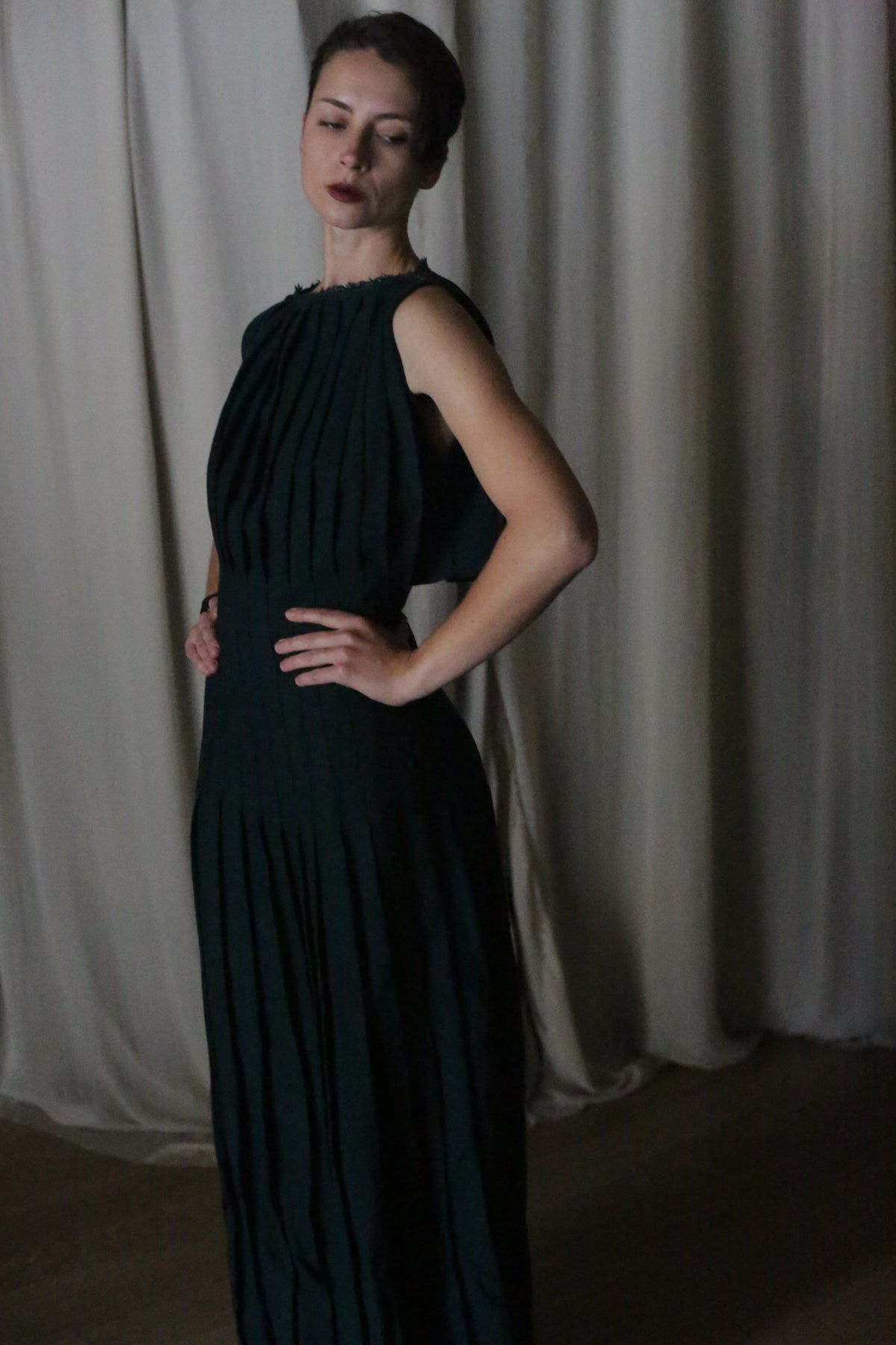 A woman with short dark hair stands with her hands on her hips, wearing the Hand Pleated Column Dress in Japanese Wool Gauze Forest, set against light-colored curtains and looking slightly down and to the side.