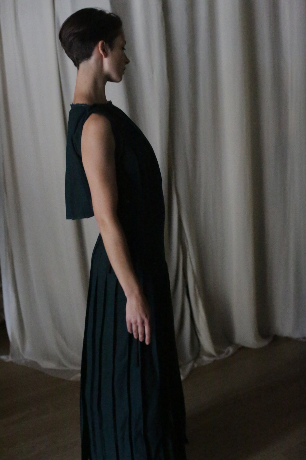 A person with short dark hair stands in profile before light curtains, wearing the Hand Pleated Column Dress in Japanese Wool Gauze Forest. Soft shadows highlight the artistry and zero-waste design of this handmade pleated garment.