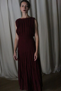 A woman stands before a curtain in a sleeveless Hand Pleated Column Dress made of Japanese wool gauze in wine. Her hair is pulled back as she gazes at the camera, with dim lighting casting soft shadows to enhance the elegance of her handmade dress.