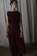 Load image into Gallery viewer, In front of a draped, cream-colored curtain, a person with short hair gazes to the side in relaxed elegance, wearing the Hand Pleated Column Dress in Japanese Wool Gauze Wine. The soft shadows cast by dim lighting highlight the handmade beauty of the sleeveless, pleated design.